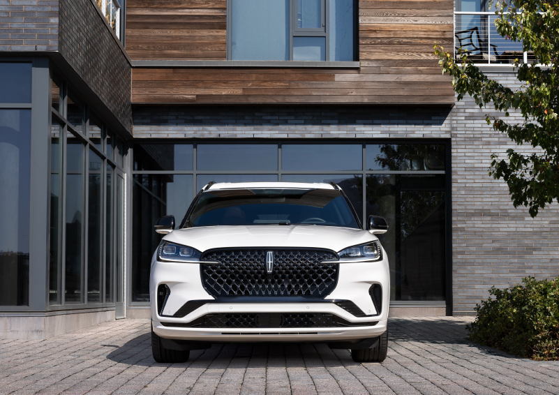 A white 2025 Lincoln Aviator® SUV is shown parked outside a modern building with gray brick and wood accents. | Oliver Lincoln in Plymouth IN