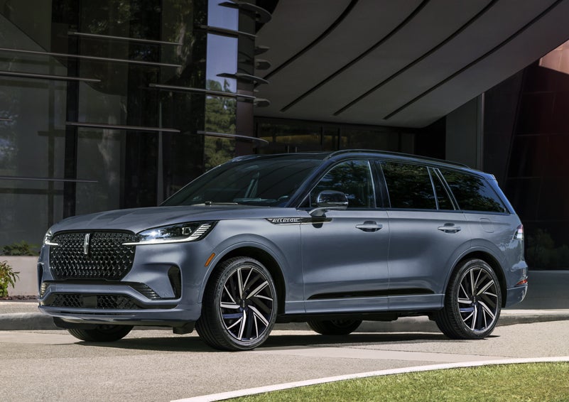 A gray 2025 Lincoln Aviator® SUV is parked outside a modern building showcasing the driver’s side of the vehicle. | Oliver Lincoln in Plymouth IN