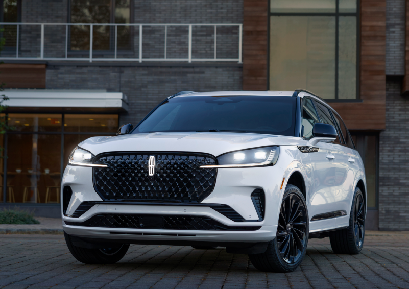 A white 2025 Lincoln Aviator® SUV is shown parked outside a modern building with gray brick and wood accents. | Oliver Lincoln in Plymouth IN