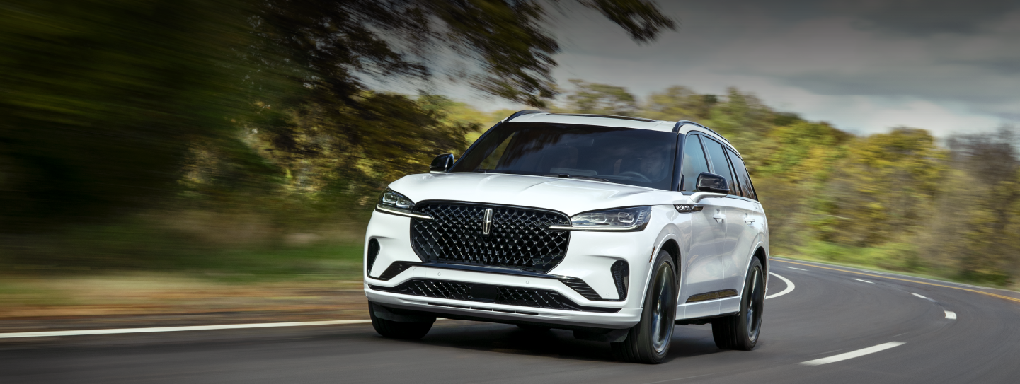 A white 2025 Lincoln Aviator® SUV is being driven down a road lined with trees. | Oliver Lincoln in Plymouth IN